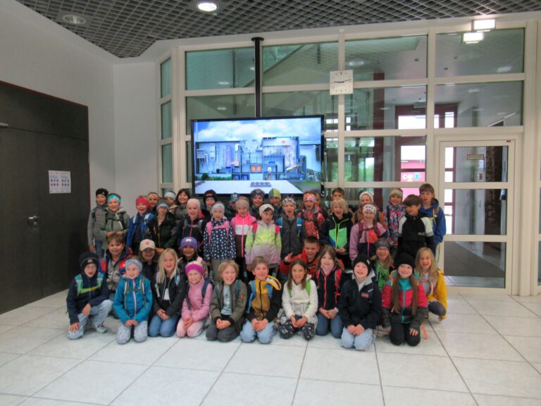 Eine gro&szlig;e Gruppe junger Kinder aus der Grundschule Grabenst&auml;tt, die Jacken und Rucks&auml;cke tragen, posieren f&uuml;r ein Gruppenfoto in einer Halle im Jahr 2026 vor einem an der Wand befestigten Bildschirm, auf dem ein Stadtbild zu sehen ist.
