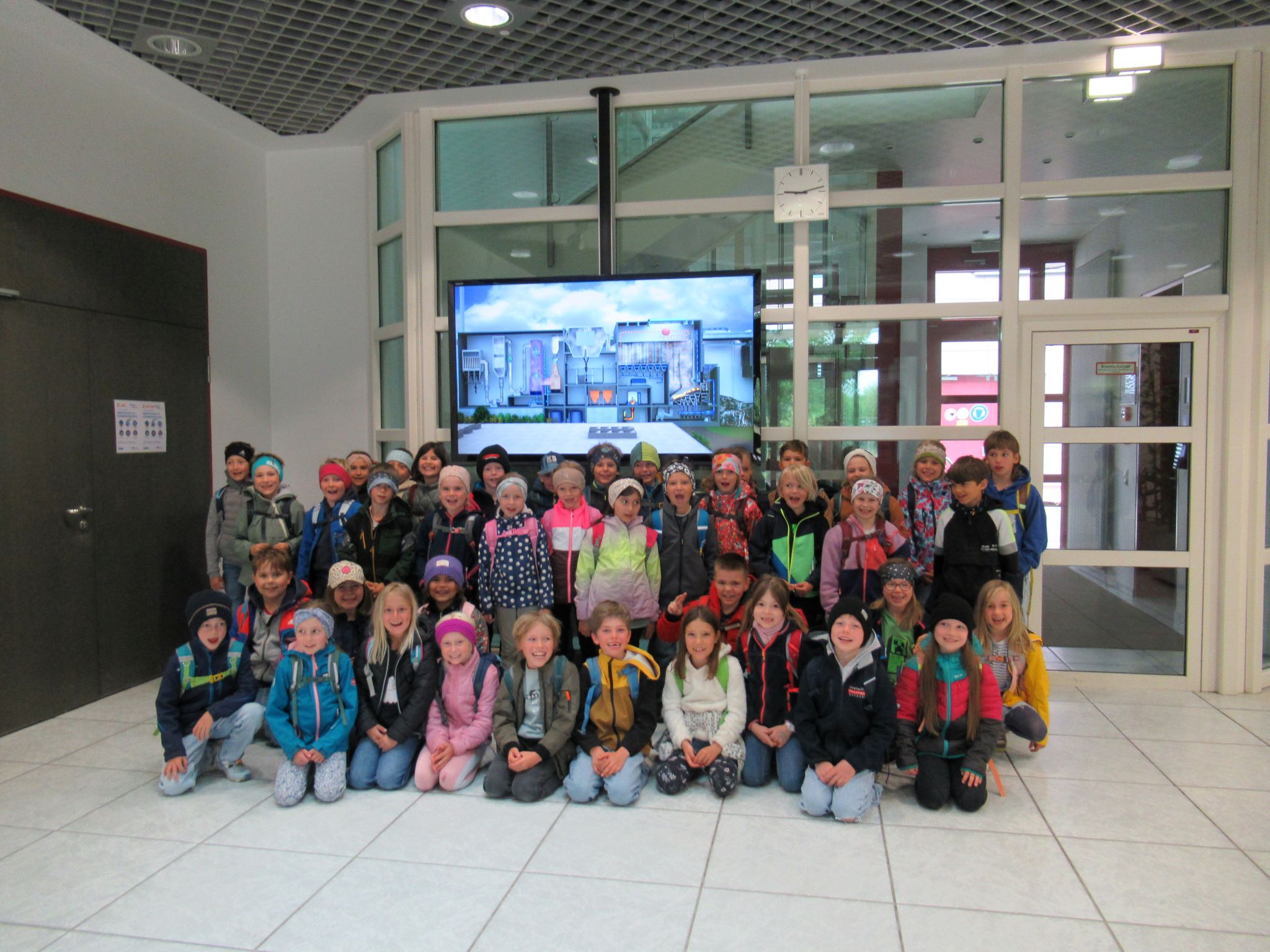 Eine gro&szlig;e Gruppe junger Kinder aus der Grundschule Grabenst&auml;tt, die Jacken und Rucks&auml;cke tragen, posieren f&uuml;r ein Gruppenfoto in einer Halle im Jahr 2026 vor einem an der Wand befestigten Bildschirm, auf dem ein Stadtbild zu sehen ist.