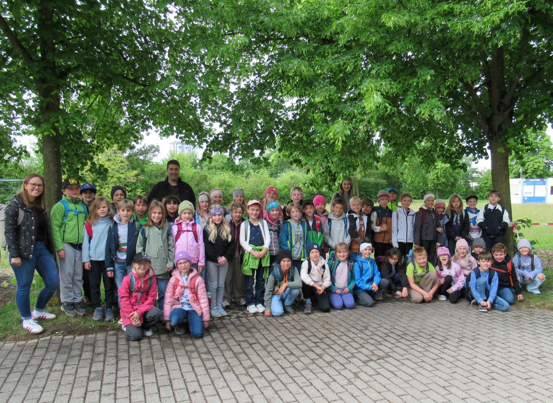 Eine gro&szlig;e Gruppe von Kindern mit Rucks&auml;cken und einige Erwachsene der Grundschule &Uuml;bersee posieren f&uuml;r ein Foto im Freien unter gr&uuml;nen B&auml;umen auf einem gepflasterten Platz, l&auml;chelnd und eng beieinander stehend am 21.05.2026.