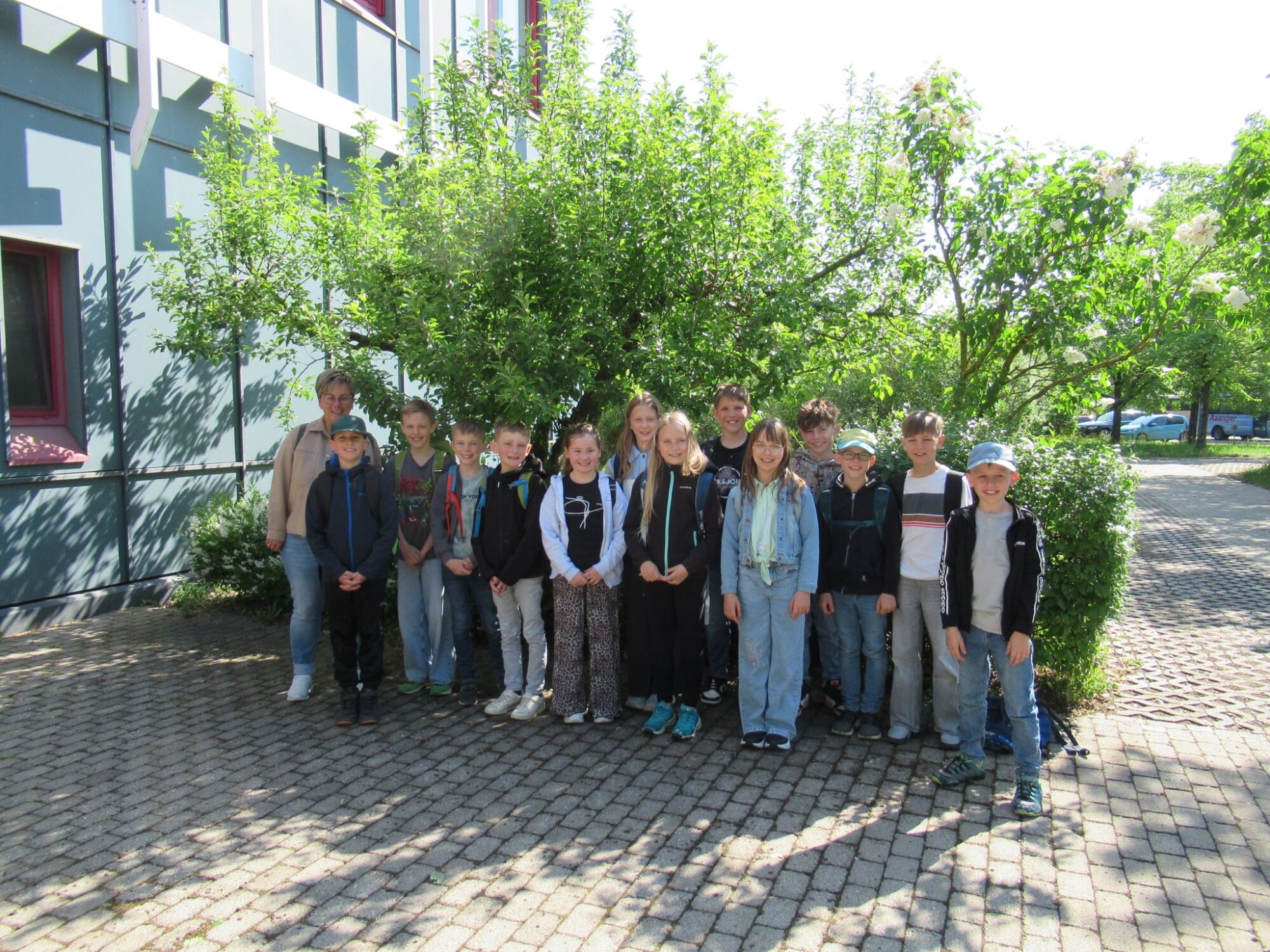 Eine Gruppe von Kindern und ein Erwachsener stehen gemeinsam drau&szlig;en auf einem gepflasterten Platz vor einem Baum und einem Geb&auml;ude und l&auml;cheln an einem sonnigen Tag in die Kamera - ein automatisch gespeicherter Entwurf eines fr&ouml;hlichen Moments.