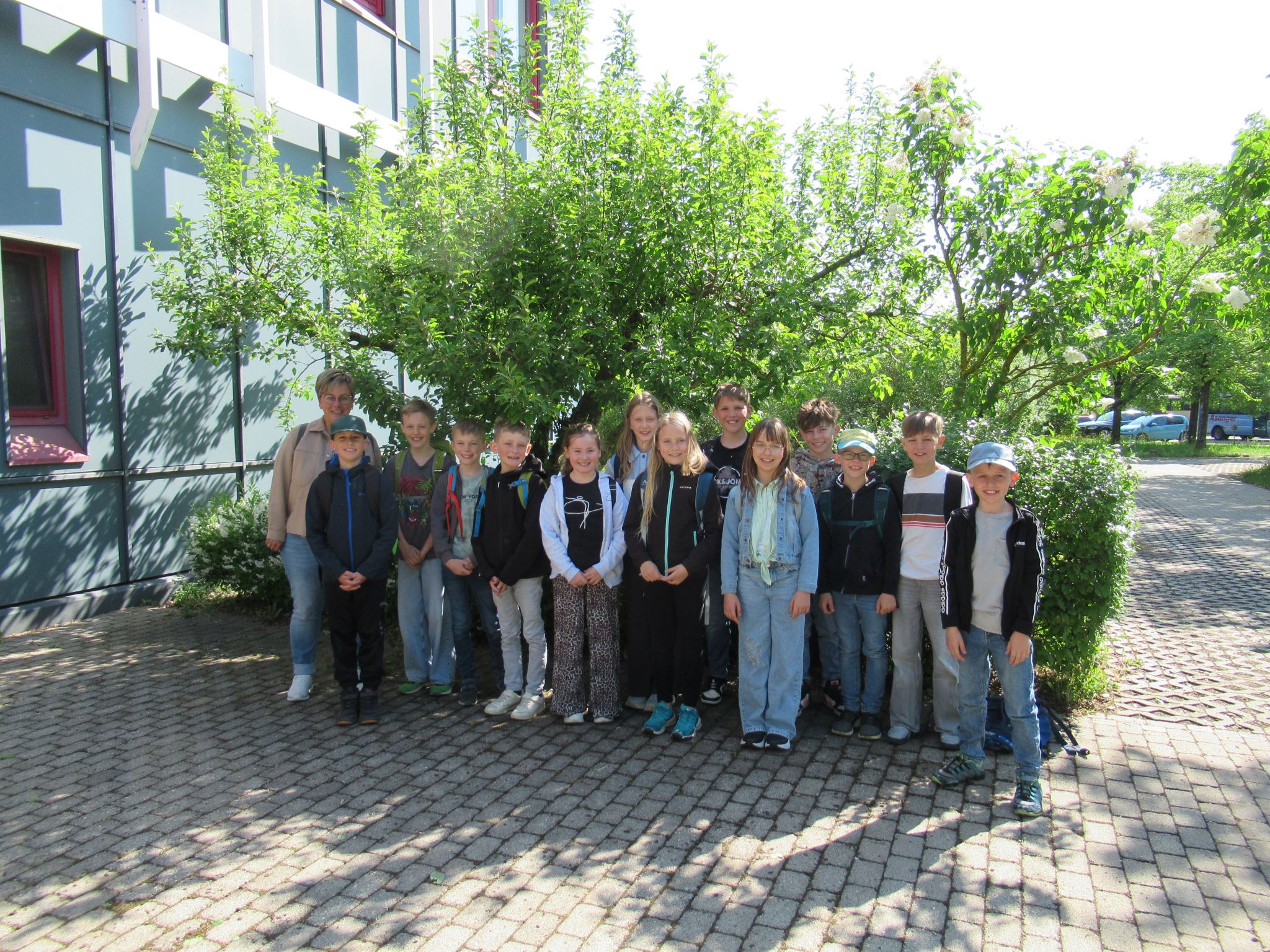 Eine Gruppe von Kindern und ein Erwachsener stehen gemeinsam drau&szlig;en auf einem gepflasterten Platz vor einem Baum und einem Geb&auml;ude und l&auml;cheln an einem sonnigen Tag in die Kamera - ein automatisch gespeicherter Entwurf eines fr&ouml;hlichen Moments.