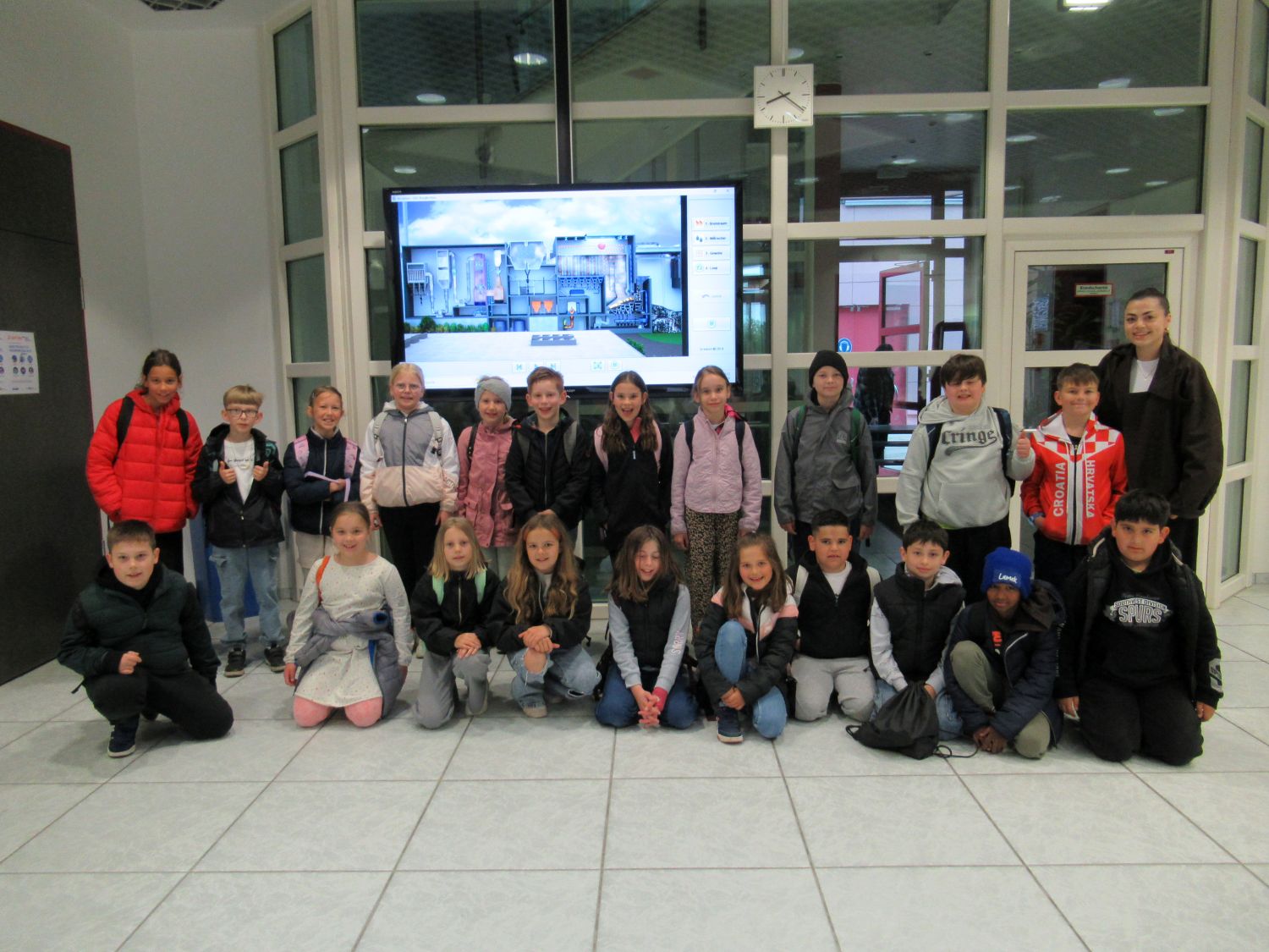 Eine Gruppe von Schulkindern und zwei Erwachsenen posiert f&uuml;r ein Foto in einem Innenraum vor einem gro&szlig;en Bildschirm und einer Uhr, stehend und kniend in zwei Reihen, alle in Freizeitkleidung und Jacken.