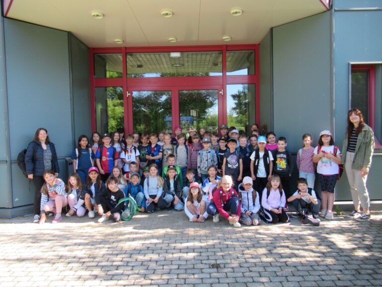 Eine gro&szlig;e Gruppe von Kindern und zwei Erwachsene posieren f&uuml;r ein Foto vor einem Geb&auml;ude mit grauen W&auml;nden und einer roten T&uuml;r. Die Kinder l&auml;cheln, einige sitzen, einige stehen, und viele tragen H&uuml;te und Rucks&auml;cke.