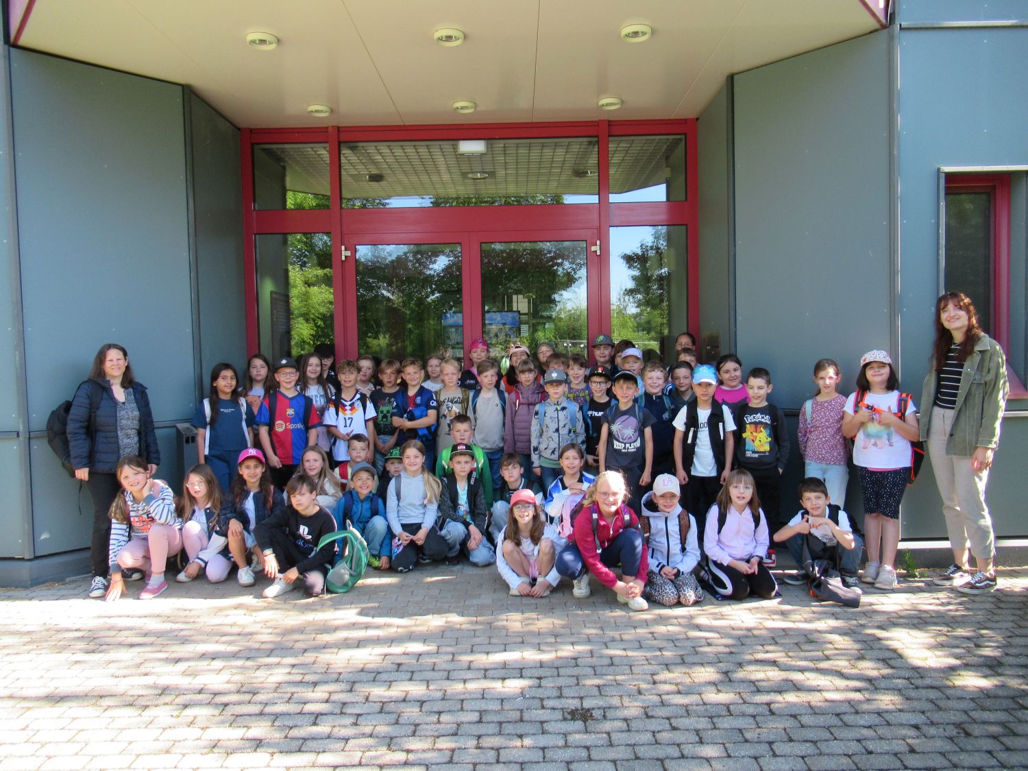 Eine gro&szlig;e Gruppe von Kindern und zwei Erwachsene posieren f&uuml;r ein Foto vor einem Geb&auml;ude mit grauen W&auml;nden und einer roten T&uuml;r. Die Kinder l&auml;cheln, einige sitzen, einige stehen, und viele tragen H&uuml;te und Rucks&auml;cke.