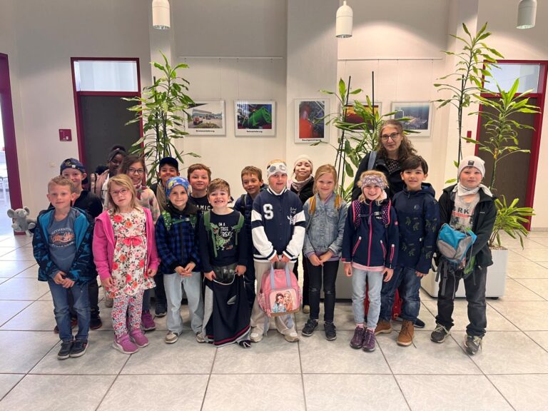 Eine Gruppe junger Kinder und ein Erwachsener stehen in zwei Reihen im Haus und l&auml;cheln in die Kamera. Sie befinden sich in einem hellen, modernen Raum mit Topfpflanzen und gerahmten Bildern an den W&auml;nden hinter ihnen.