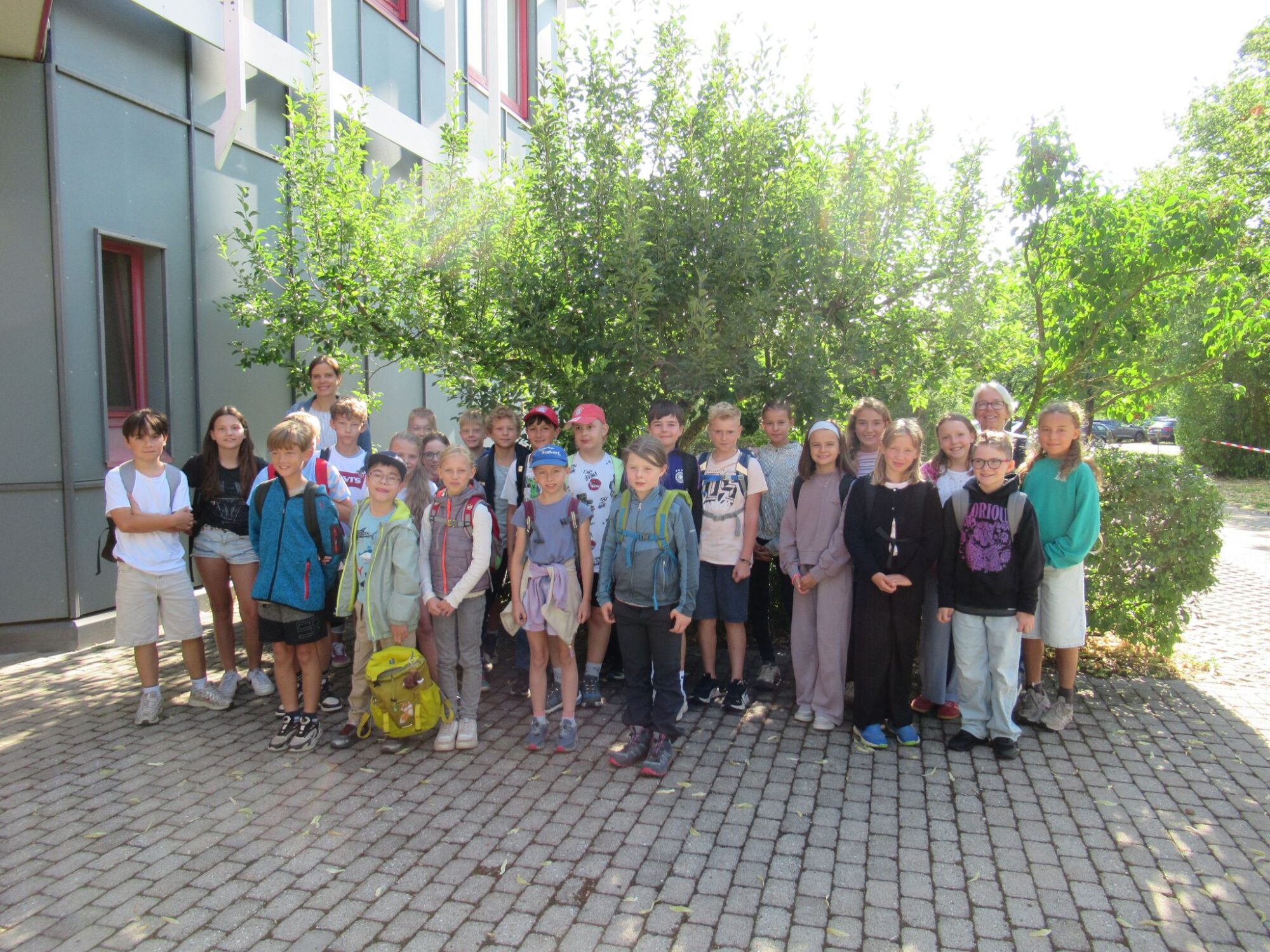 Eine Gruppe von Kindern und zwei Erwachsene stehen gemeinsam auf einem Kopfsteinpflasterweg vor einem Geb&auml;ude, im Hintergrund sind gr&uuml;ne B&auml;ume und Str&auml;ucher zu sehen. Die meisten Kinder, bei denen es sich offenbar um Sch&uuml;ler der Grundschule Surberg handelt, tragen Rucks&auml;cke und l&auml;cheln in die Kamera. Das Foto wurde am 09.07.2026 aufgenommen.