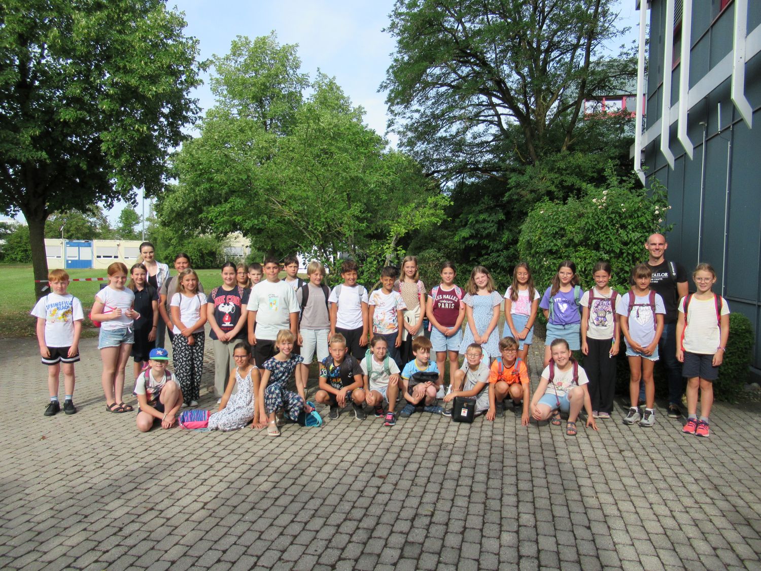Eine gro&szlig;e Gruppe von Kindern und zwei Erwachsene posieren drau&szlig;en auf einer gepflasterten Fl&auml;che, die von gr&uuml;nen B&auml;umen und Str&auml;uchern umgeben ist, f&uuml;r ein Foto. Die Kinder tragen Sommerkleidung, und einige haben Rucks&auml;cke dabei.