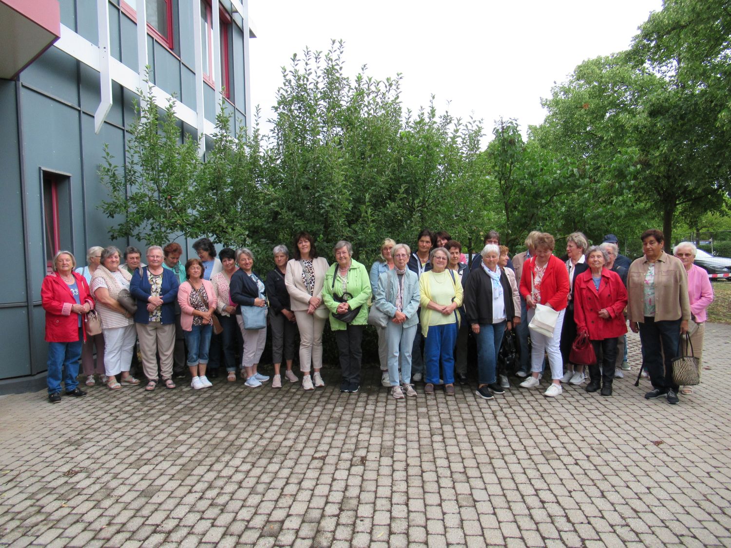 Eine Gruppe von etwa 25 Personen, &uuml;berwiegend &auml;ltere Menschen, steht gemeinsam auf einer gepflasterten Fl&auml;che im Freien in der N&auml;he eines modernen Geb&auml;udes und &uuml;ppig gr&uuml;ner B&auml;ume und posiert f&uuml;r ein Gruppenfoto.