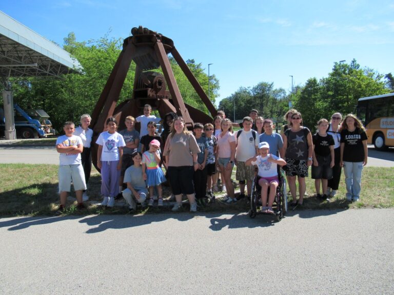 Eine Gruppe von Menschen, darunter Kinder und Erwachsene, von denen einige eine Behinderung haben, posiert an einem sonnigen Tag gemeinsam vor einer gro&szlig;en Metallglocke. Im Hintergrund sind ein Bus und B&auml;ume zu sehen.