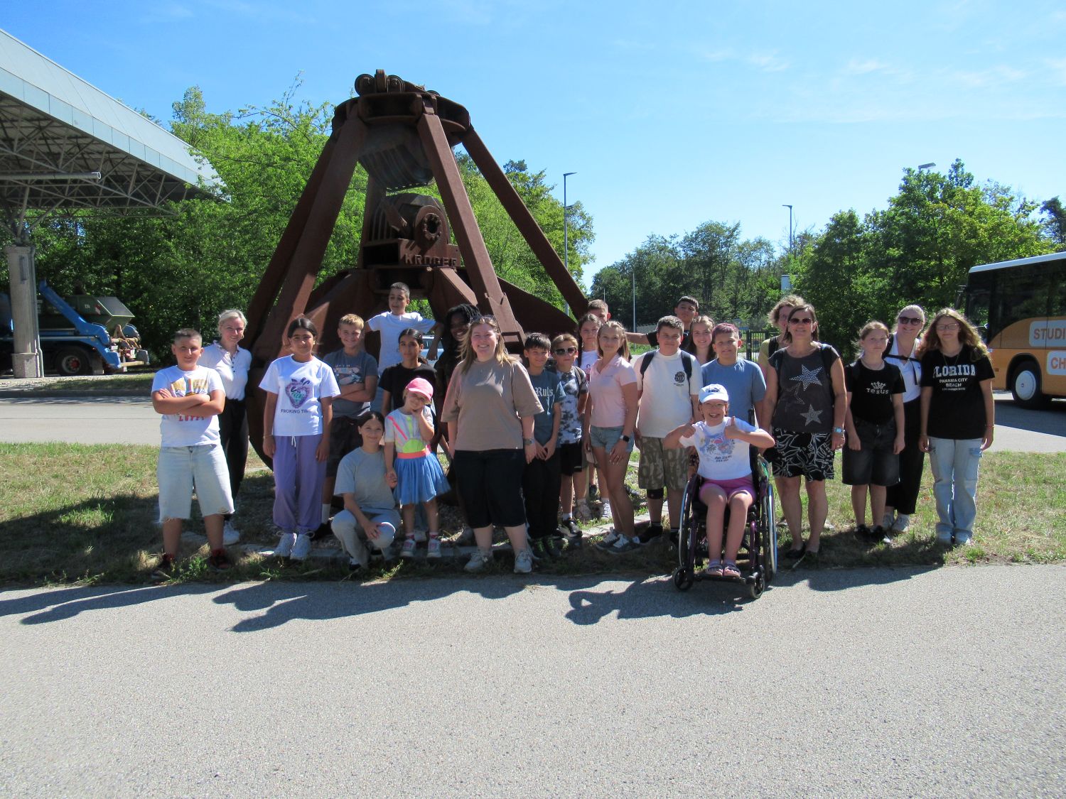 Eine Gruppe von Menschen, darunter Kinder und Erwachsene, von denen einige eine Behinderung haben, posiert an einem sonnigen Tag gemeinsam vor einer gro&szlig;en Metallglocke. Im Hintergrund sind ein Bus und B&auml;ume zu sehen.