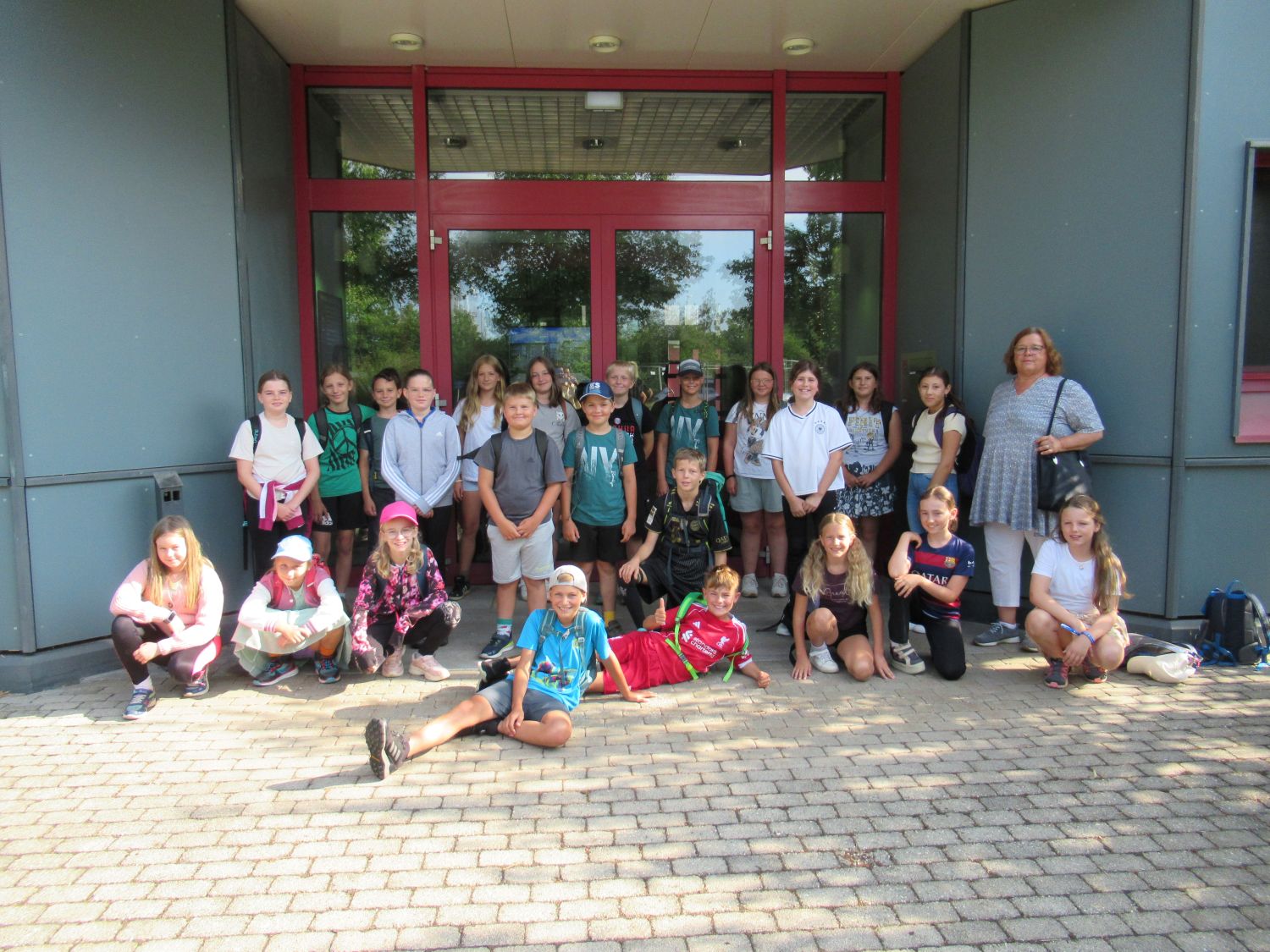 Eine Gruppe von Kindern und ein Erwachsener posieren vor einem Geb&auml;ude mit rot gerahmten Glast&uuml;ren f&uuml;r ein Foto. Die meisten stehen, einige sitzen oder knien auf dem Boden, und alle scheinen zu l&auml;cheln.