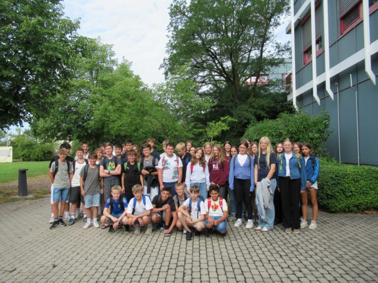 Eine Gruppe von Sch&uuml;lern posiert gemeinsam drau&szlig;en auf einer gepflasterten Fl&auml;che, im Hintergrund sind Gr&uuml;nfl&auml;chen und B&auml;ume zu sehen, direkt neben einem modernen Geb&auml;ude.