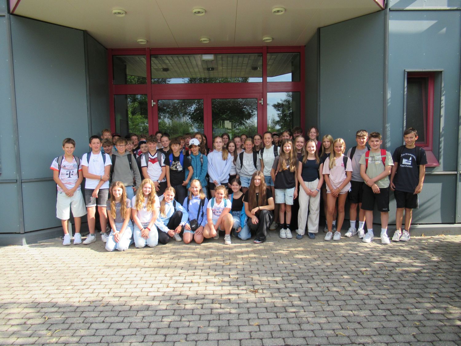 Eine Gruppe von Sch&uuml;lern posiert vor einem Schulgeb&auml;ude f&uuml;r ein Foto; sie stehen und sitzen auf einem gepflasterten Gehweg vor Glast&uuml;ren.