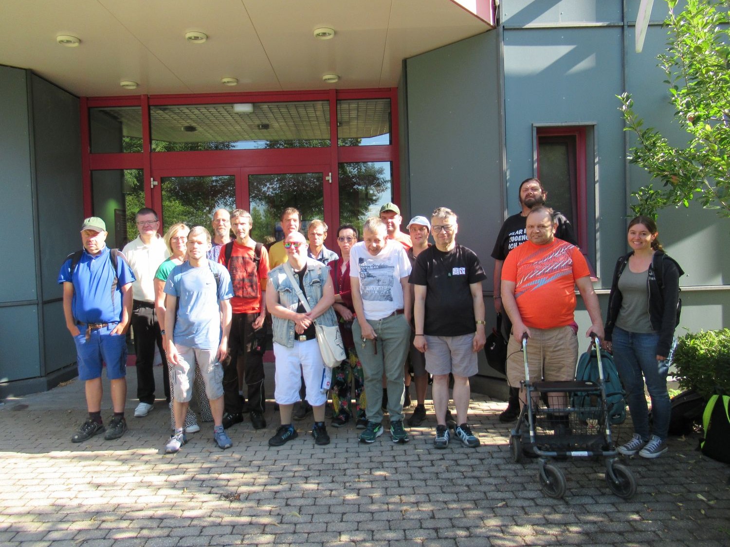 Eine Gruppe von Menschen steht l&auml;chelnd vor dem Eingang eines Geb&auml;udes; einige halten Wanderst&ouml;cke in der Hand oder st&uuml;tzen sich auf einen Rollator. Die Gruppe ist leger gekleidet, um sie herum sind Gr&uuml;nfl&auml;chen und Sonnenlicht zu sehen.