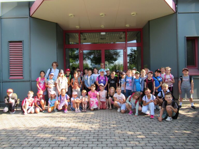 Eine Gruppe von Kindern und zwei Erwachsene posieren gemeinsam vor einem Geb&auml;ude mit Glast&uuml;ren und roten Zierleisten; sie stehen und hocken auf einem gepflasterten Weg. Die Kinder tragen legere Sommerkleidung und Rucks&auml;cke.