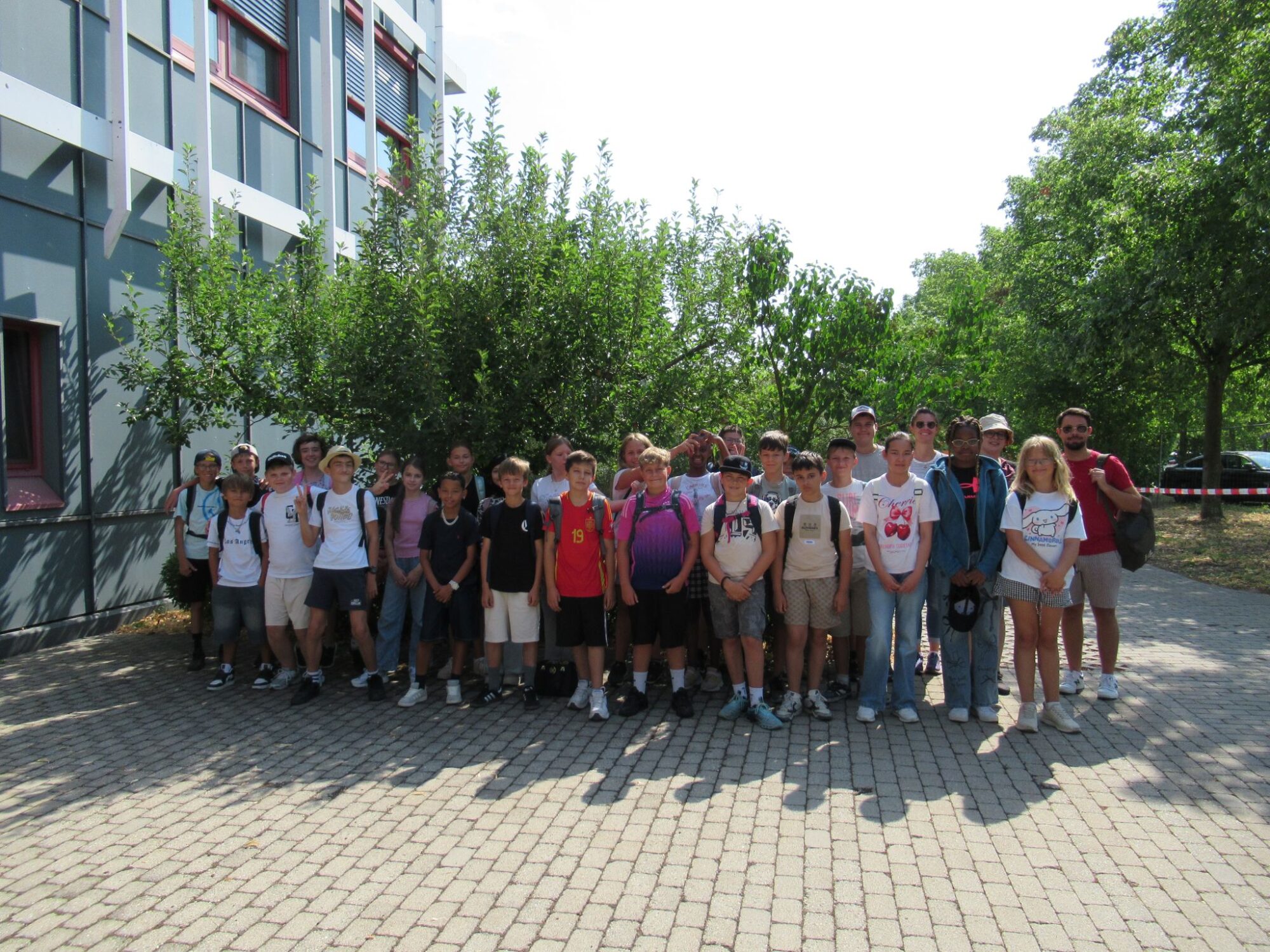 Eine Gruppe von Sch&uuml;lern und Erwachsenen steht an einem sonnigen Tag gemeinsam im Freien und posiert f&uuml;r ein Gruppenfoto vor einem Geb&auml;ude und einigen B&auml;umen an der Mittelschule M&uuml;hldorf. Die meisten l&auml;cheln und tragen Freizeitkleidung sowie Rucks&auml;cke.
