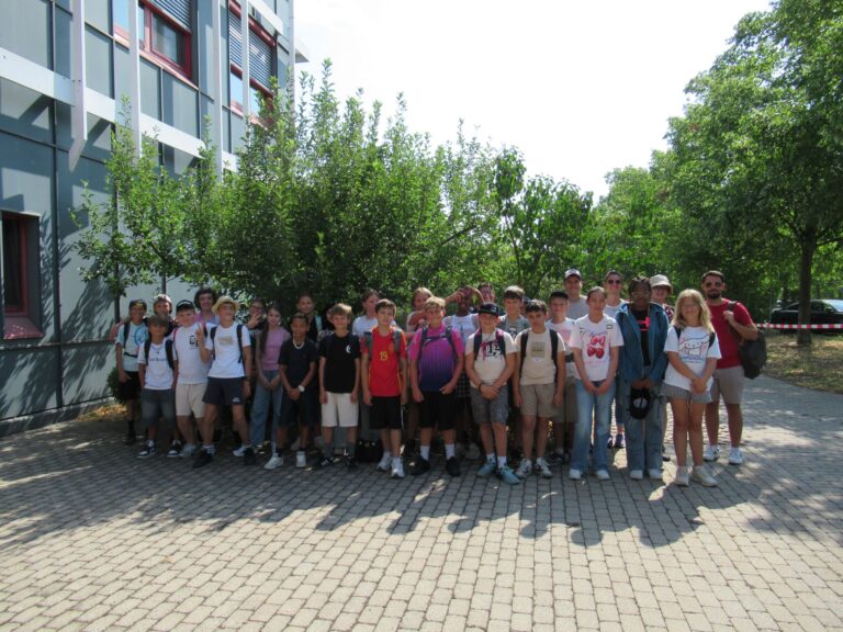 Eine Gruppe von Sch&uuml;lern und Erwachsenen steht an einem sonnigen Tag gemeinsam im Freien und posiert f&uuml;r ein Gruppenfoto vor einem Geb&auml;ude und einigen B&auml;umen an der Mittelschule M&uuml;hldorf. Die meisten l&auml;cheln und tragen Freizeitkleidung sowie Rucks&auml;cke.