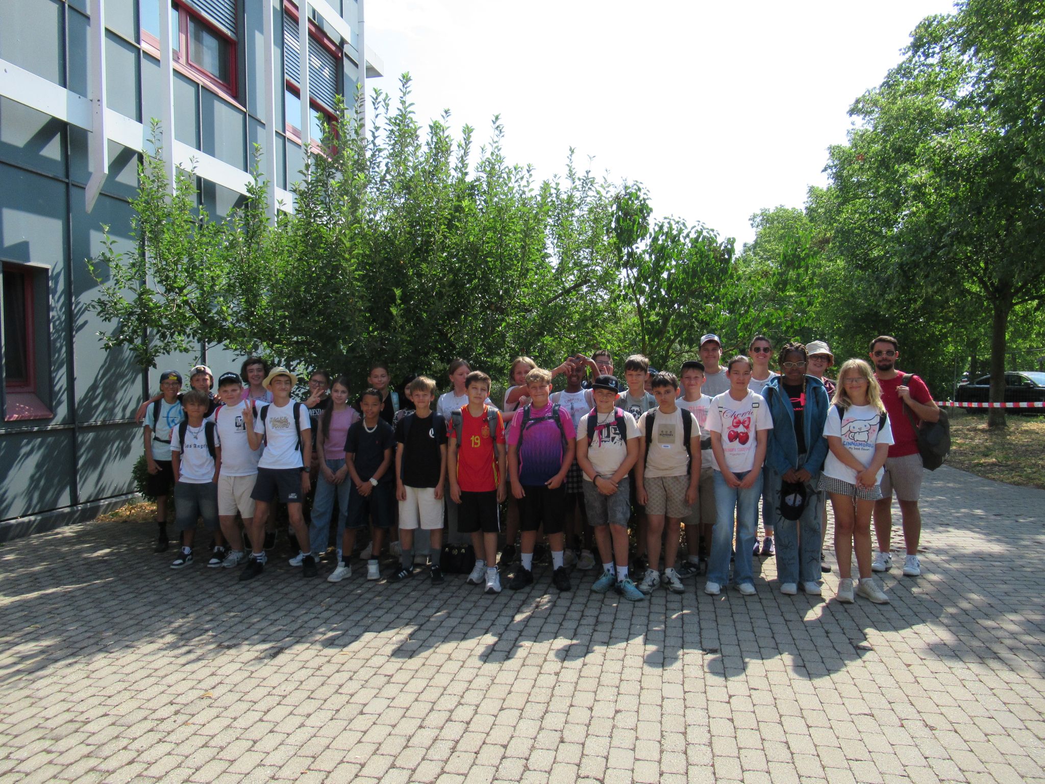 Eine Gruppe von Sch&uuml;lern und Erwachsenen steht an einem sonnigen Tag gemeinsam im Freien und posiert f&uuml;r ein Gruppenfoto vor einem Geb&auml;ude und einigen B&auml;umen an der Mittelschule M&uuml;hldorf. Die meisten l&auml;cheln und tragen Freizeitkleidung sowie Rucks&auml;cke.