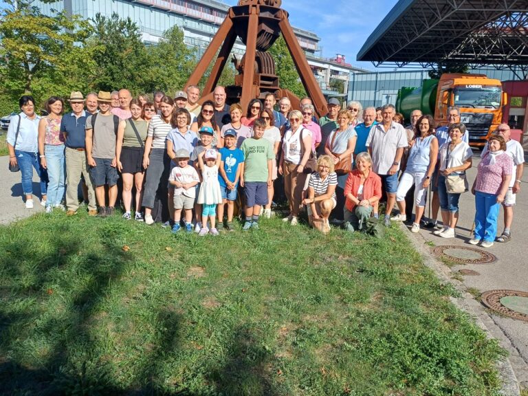 Eine Gruppe von Erwachsenen und Kindern posiert an einem sonnigen Tag gemeinsam im Freien vor einer gro&szlig;en Industrieskulptur, im Hintergrund sind Geb&auml;ude, B&auml;ume und ein orangefarbener Lkw zu sehen.