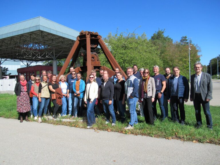 Eine Gruppe von etwa 25 Erwachsenen, die zum Kollegium der Berufsschule Alt&ouml;tting geh&ouml;ren, steht l&auml;chelnd im Freien vor einer gro&szlig;en industriellen Metallkonstruktion. Im Hintergrund sind unter einem strahlend blauen Himmel B&auml;ume und ein Geb&auml;ude mit schr&auml;gem Dach zu sehen.