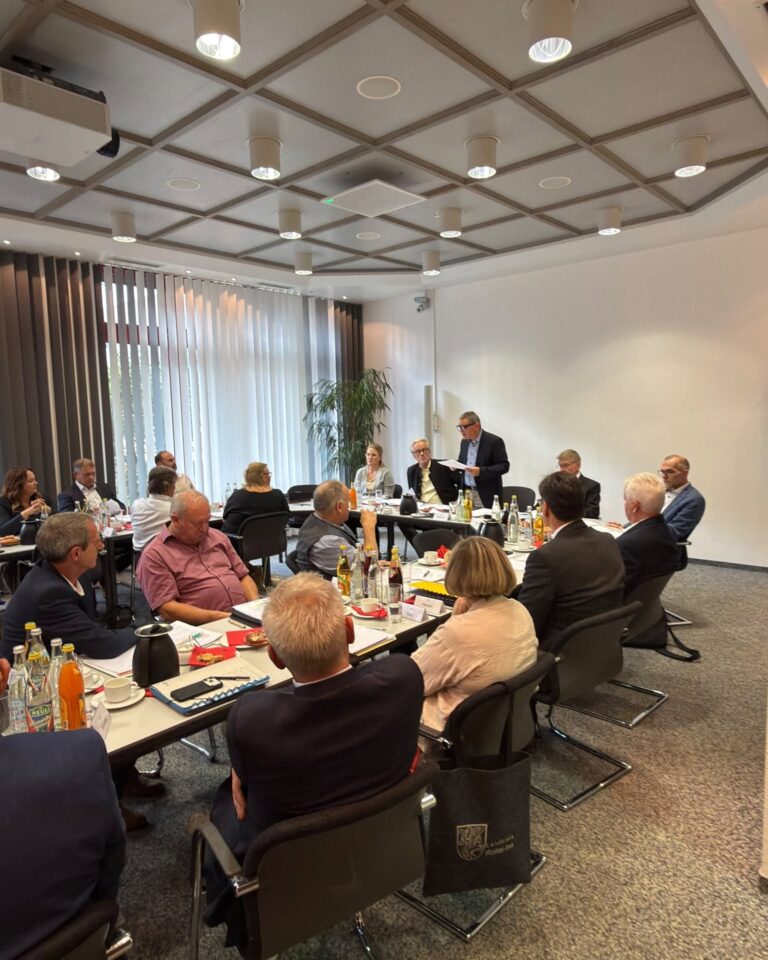 Verbandsvorsitzender Erwin Schneider steht am Kopfende eines Konferenztisches und spricht zu einer Gruppe von Personen, die zu einer Besprechung in einem Sitzungssaal Platz genommen haben.