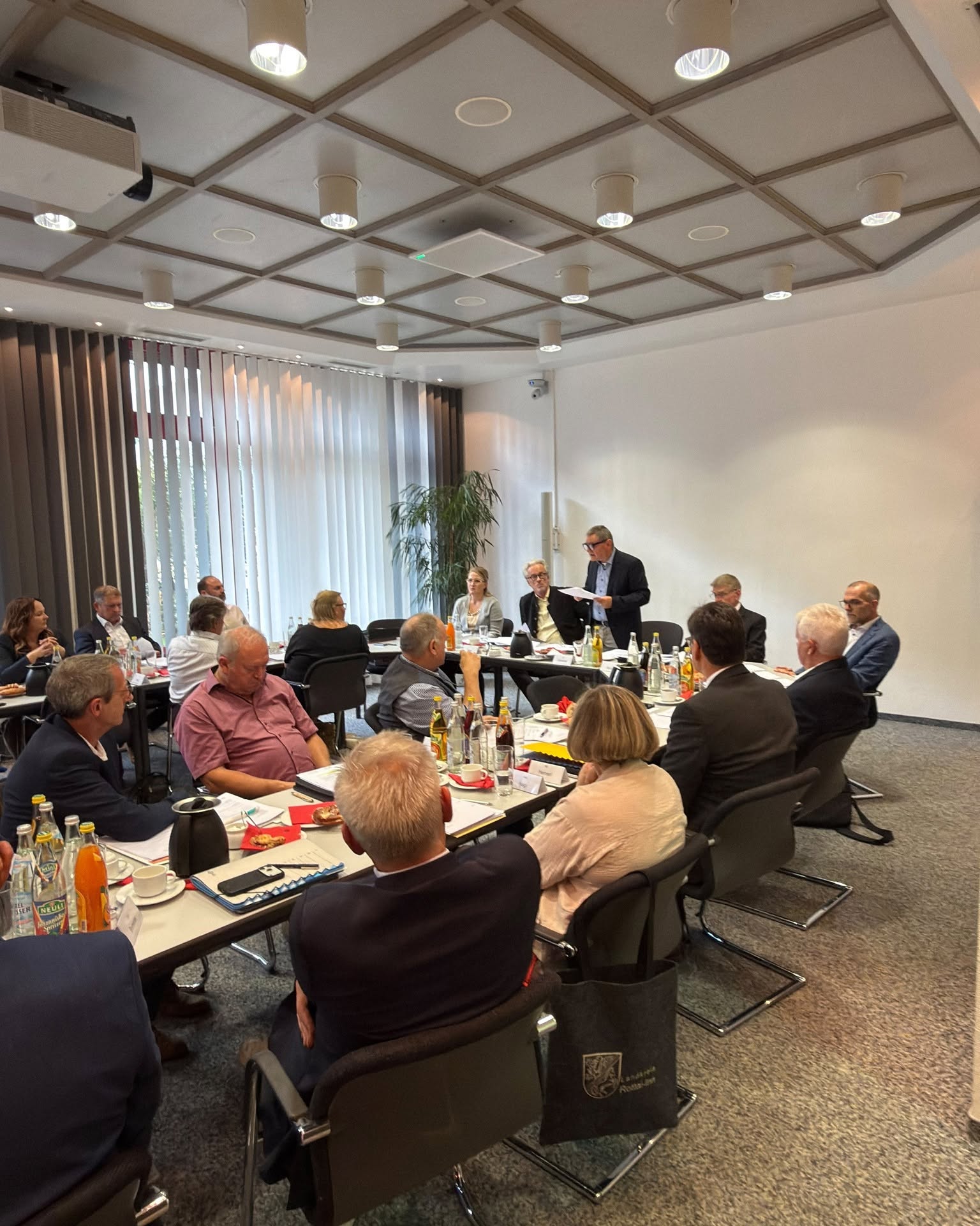 Verbandsvorsitzender Erwin Schneider steht am Kopfende eines Konferenztisches und spricht zu einer Gruppe von Personen, die zu einer Besprechung in einem Sitzungssaal Platz genommen haben.